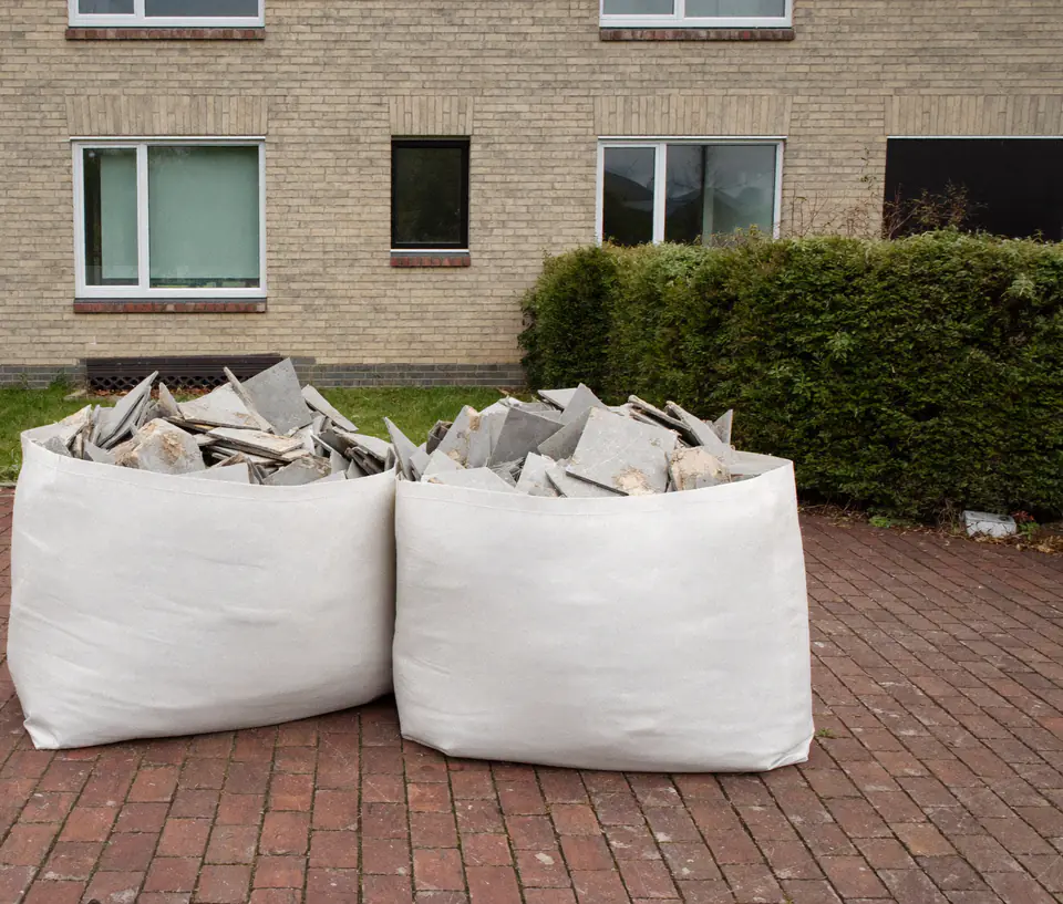 Twee gevulde big bags met gebroken grijze tegels op een bestrate oprit voor een woning met een lichte bakstenen gevel